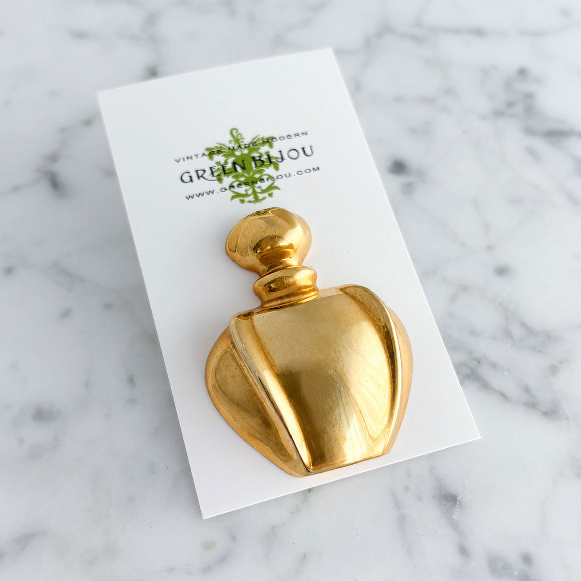 Polished gold-tone perfume bottle brooch pinned to a cream ribbed turtleneck