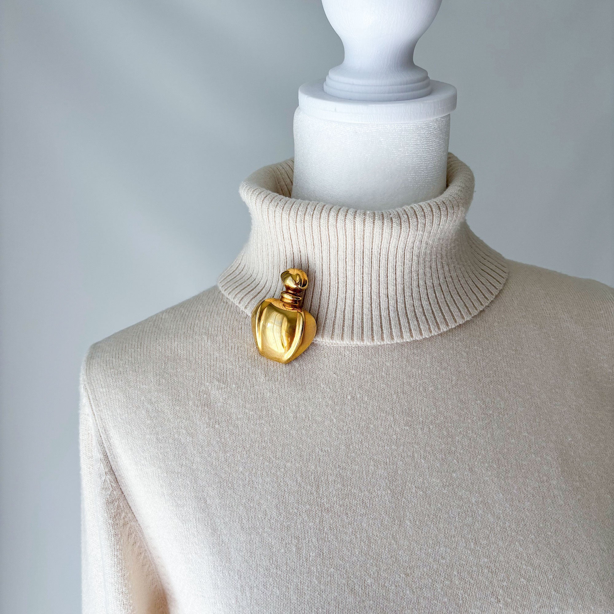 Polished gold-tone perfume bottle brooch pinned to a cream ribbed turtleneck
