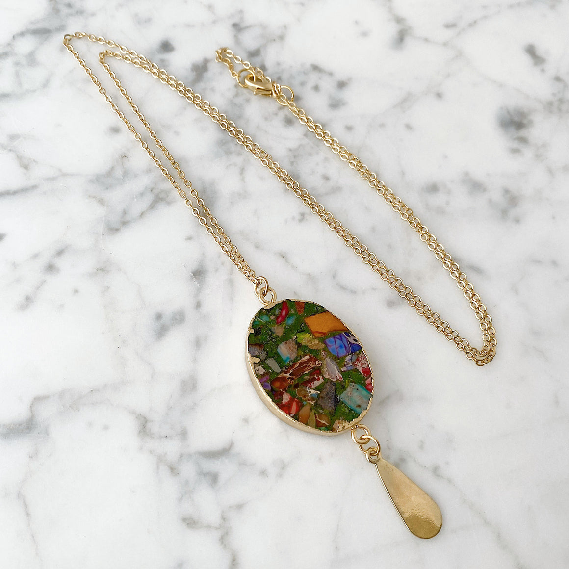 Close-up of a multicolour oval jasper pendant with gold-tone edging and a dangling gold-tone teardrop charm on white marble