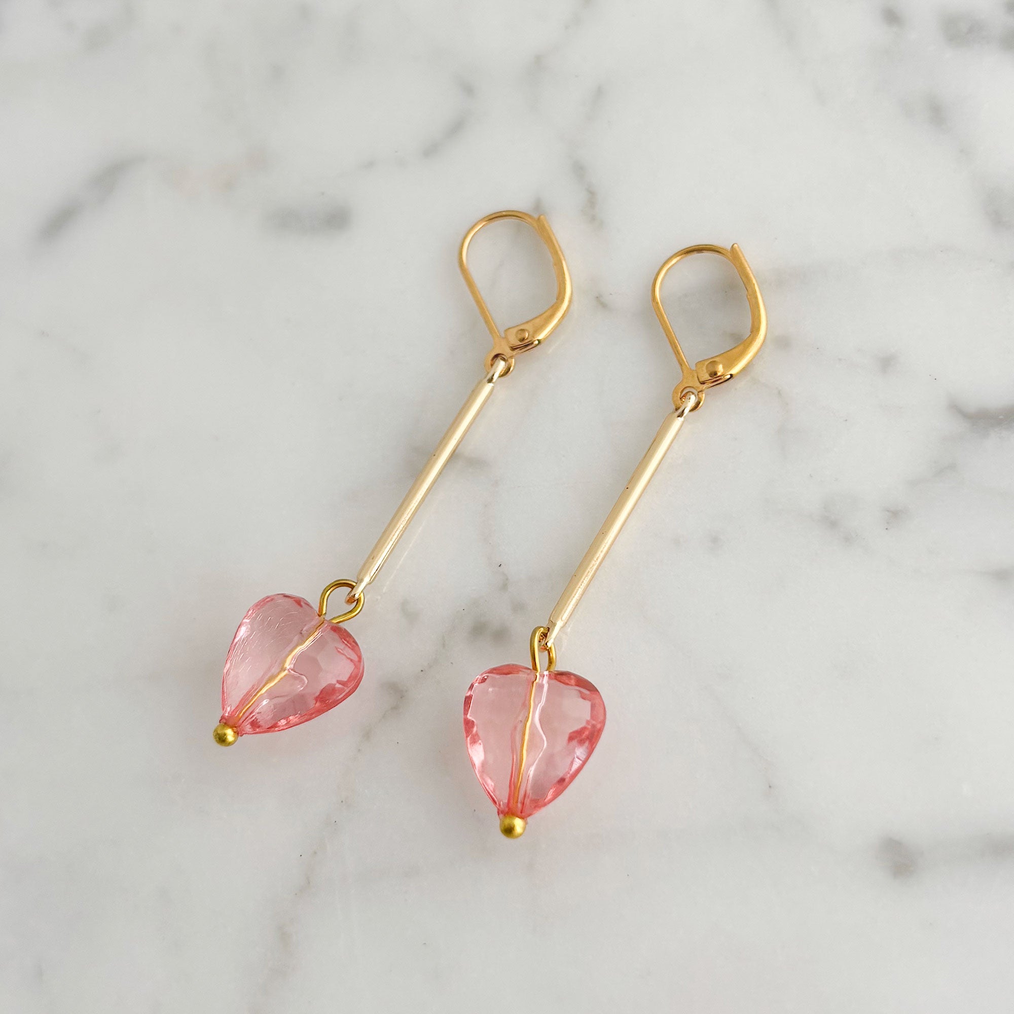 Pair of faceted translucent pink heart earrings with long gold-tone bars on white marble