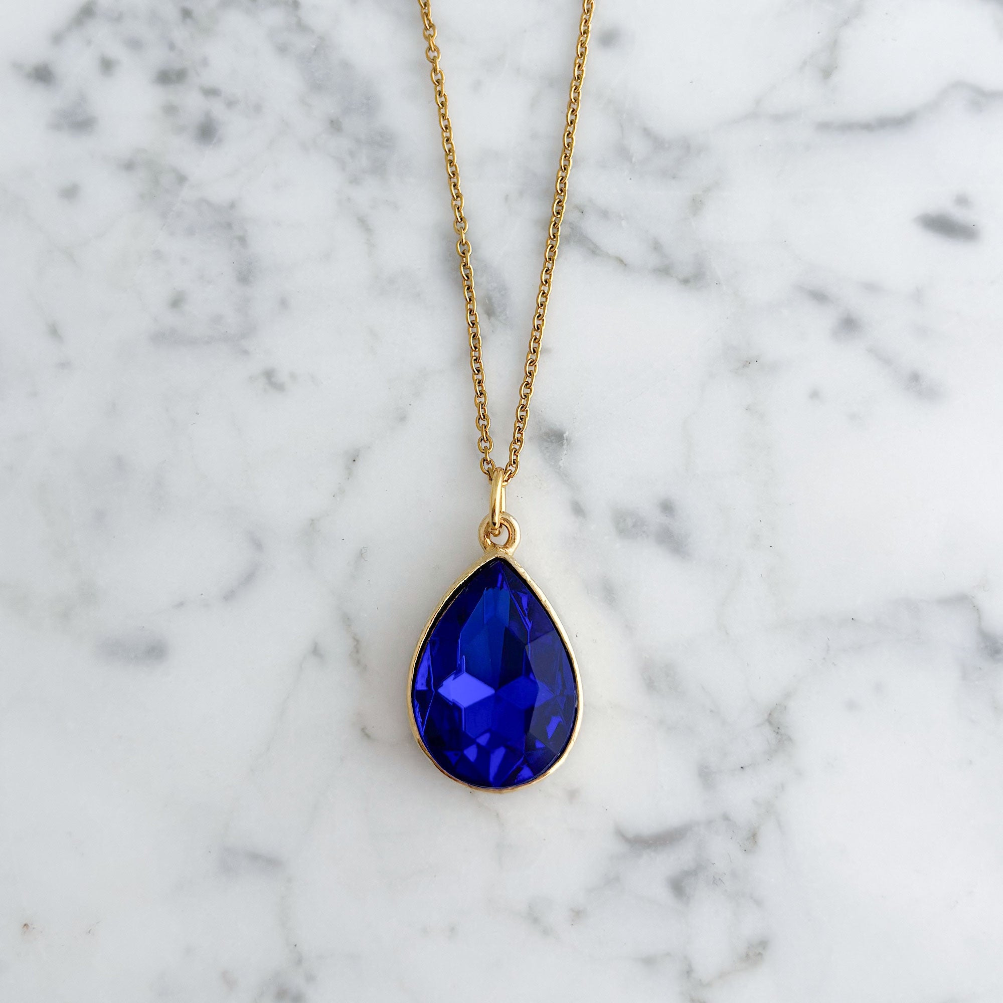 Deep blue faceted teardrop crystal in a gold-tone setting on white marble
