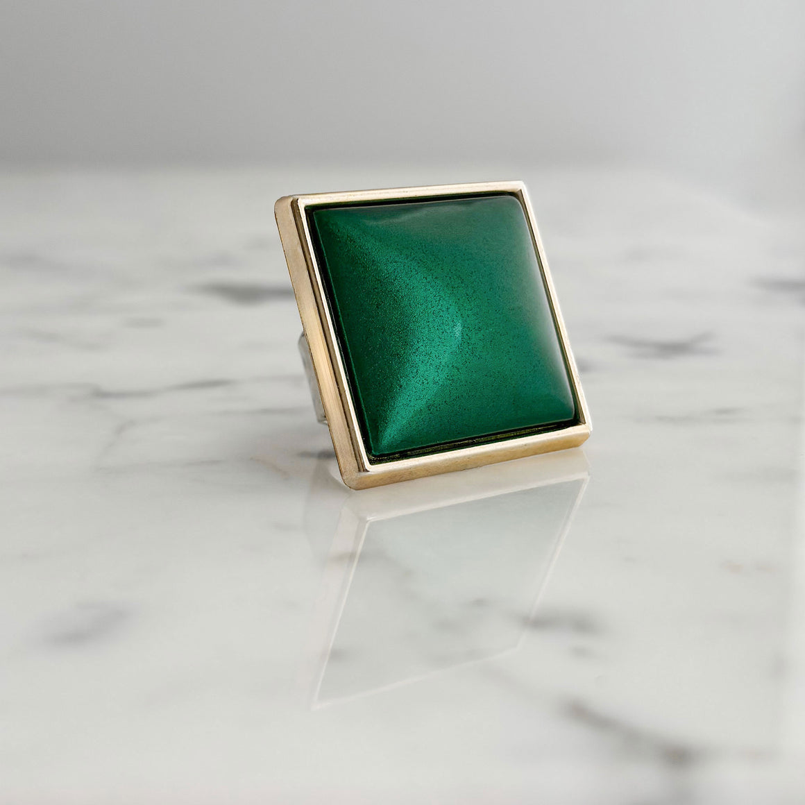 Hand wearing a large square emerald-green ring with a domed pearlescent centre and gold-tone frame