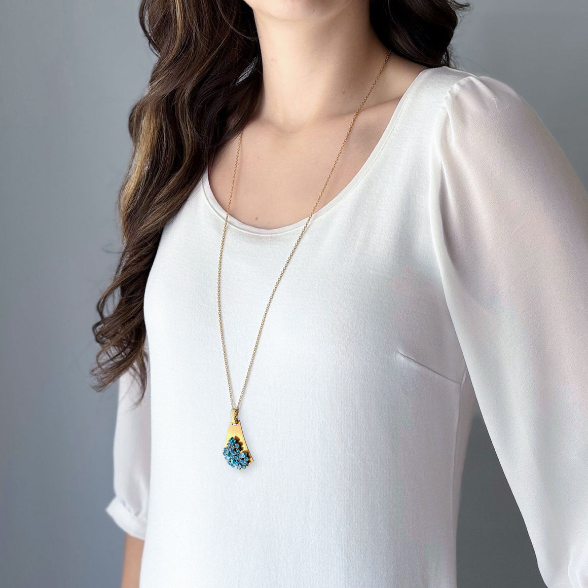 Close-up of a gold-tone shield-shaped pendant with a floral cluster of sparkling teal rhinestones on white marble
